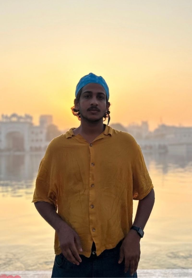 A young man in a mustard-yellow shirt and a blue head wrap standing at the edge of still water in low golden light, a pale domed building faint in the haze behind him.